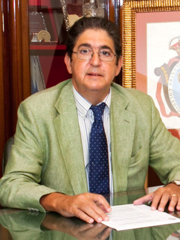 Hombre en traje de abogado sentado en un escritorio con documentos, en un ambiente formal con escudo del Colegio de Abogados de fondo.
