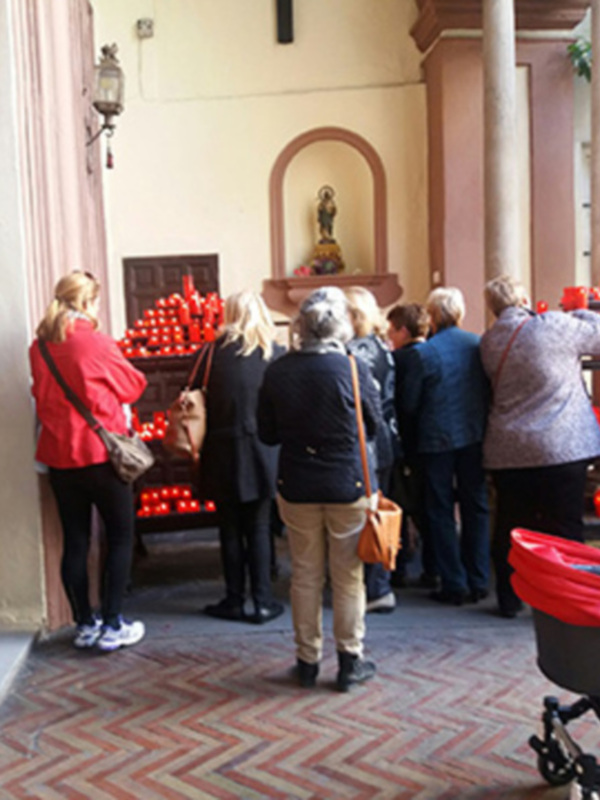 Gente rezando frente a una capilla con velas rojas en un patio interior.