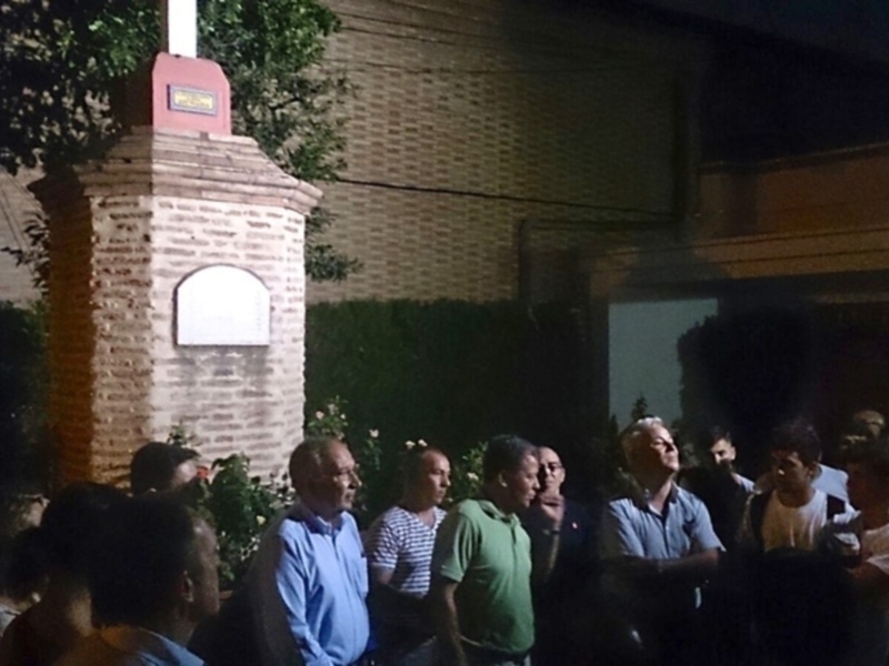 Grupo de personas reunidas en una plaza nocturna, cerca de un monumento con una placa en la pared de ladrillo.
