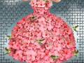 Una mujer con un vestido hecho de flores rosadas, posa en una plataforma de madera. El fondo es un muro de rejilla metálica y césped verde.