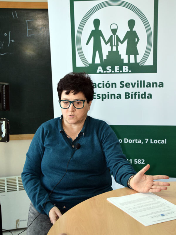 A una persona sentada en un escritorio, con un fondo que incluye un cartel de la Asociación Sevillana de Espina Bífida (ASEB). La persona está hablando y gesturando con una mano. La mesa tiene un papel en blanco sobre ella, y el fondo incluye un tablero de madera con letras escritas.