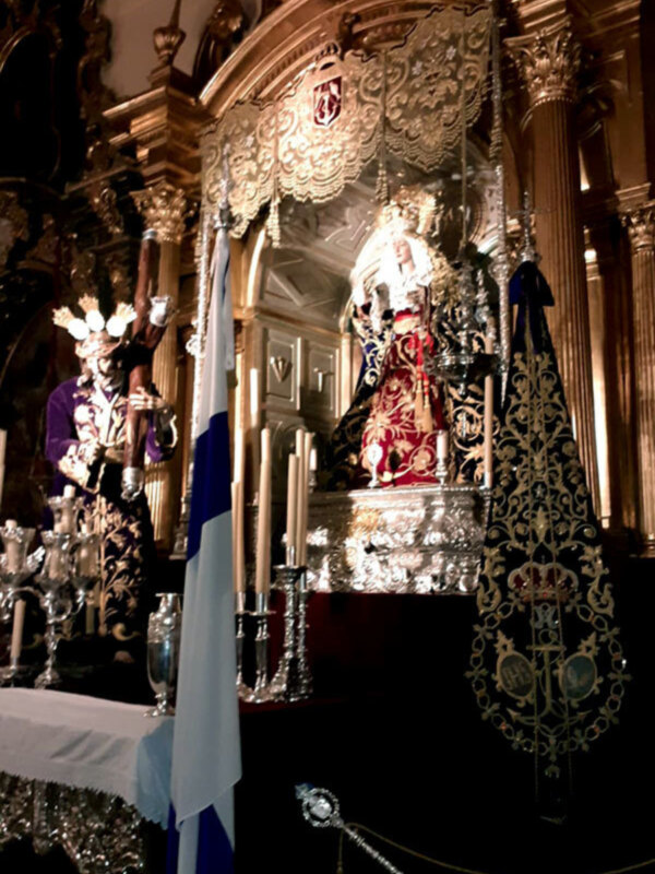 Procesión religiosa con estandarte y figura procesional en un entorno ornamental y candelabros de plata.