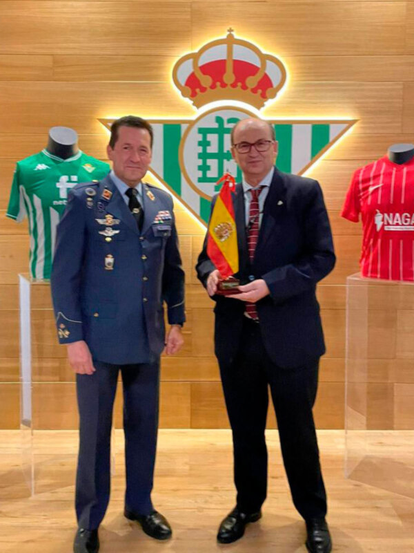 Dos hombres posan con una medalla y dos camisetas de fútbol en un contexto formal.