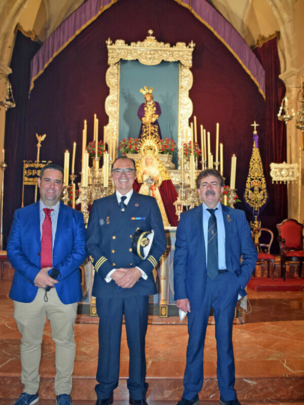 Tres hombres en trajes formales posan frente a una imagen religiosa adornada con flores y velas en un interior ornamental.