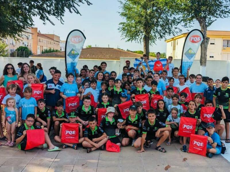Equipo de jóvenes atletas posando con banderas y bolsas de PQS en un evento deportivo al aire libre.