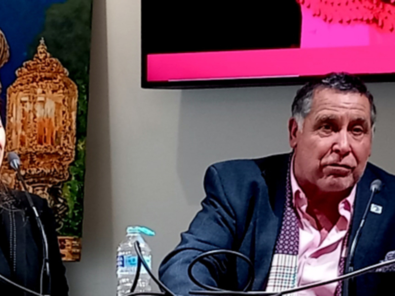 Persona hablando en un evento público con un fondo de imagen religiosa y un monitor de pantalla en el fondo.