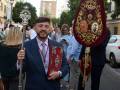 Un hombre en traje sostiene un bastón ceremonial con un escudo heráldico y una cruz en la punta, durante una procesión o ceremonia formal.