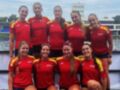 Equipo femenino de remo posando en una cancha deportiva con el agua al fondo.