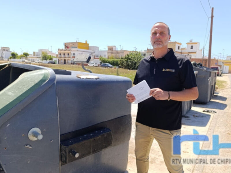 Un hombre sostiene un papel junto a una papelería, posiblemente en un entorno urbano con edificios al fondo.
