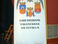 Un cartel blanco con un escudo de armas y el texto "Casa Regional Valenciana en Sevilla" en una sala con asientos.