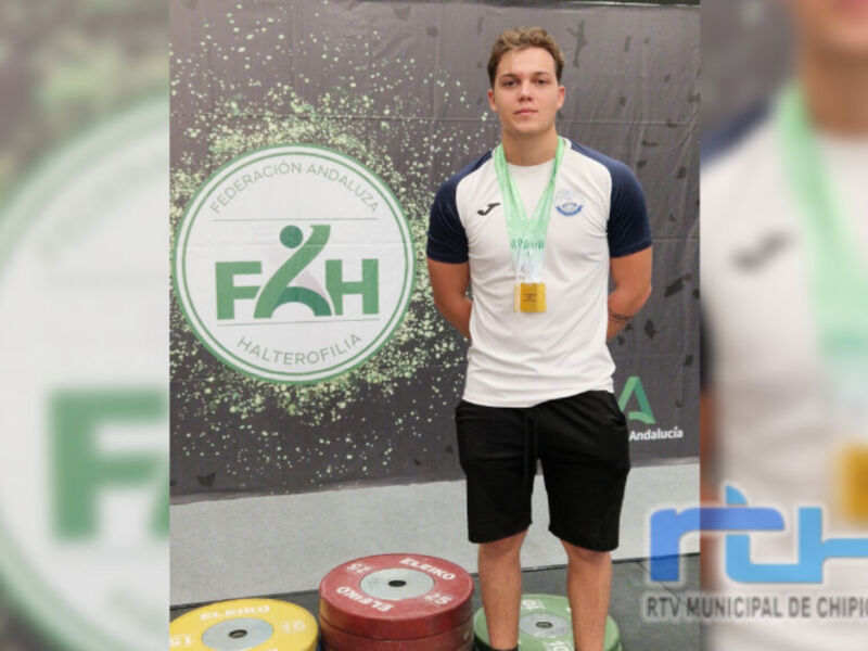 Un atleta vestido con una camiseta blanca y pantalones cortos negros, posando orgulloso con una medalla dorada al cuello. Fondo con un banner de la Federación Andaluza de Halterofilia (FAHH) y letras en verde.
