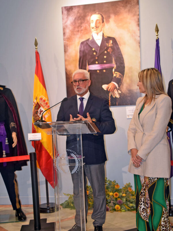 Un hombre y una mujer en trajes formales se encuentran frente a un escenario con un micrófono y una bandera española. En el fondo, hay un retrato de un hombre en uniforme militar y flores en la alfombra.
