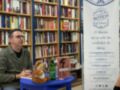 Un hombre con gafas sentado en una silla azul frente a estanterías de libros. En la mesa hay un libro abierto y una figura de peluche. A su lado, se encuentra un cartel publicitario que anuncia la librería Botica de Lecturas.