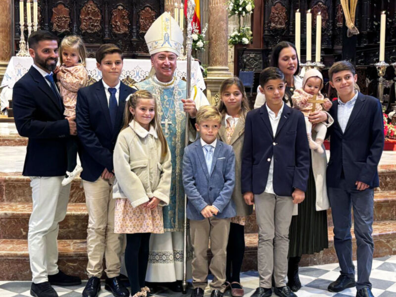 En una celebración religiosa, un sacerdote y varios niños posan en la escalinata de una iglesia. El sacerdote, vestido con un hábito blanco y dorado, sostiene una cruz. Los niños están vestidos formalmente, con chaquetas y faldas. La imagen muestra una escena de fe y familia en un ambiente histórico.
