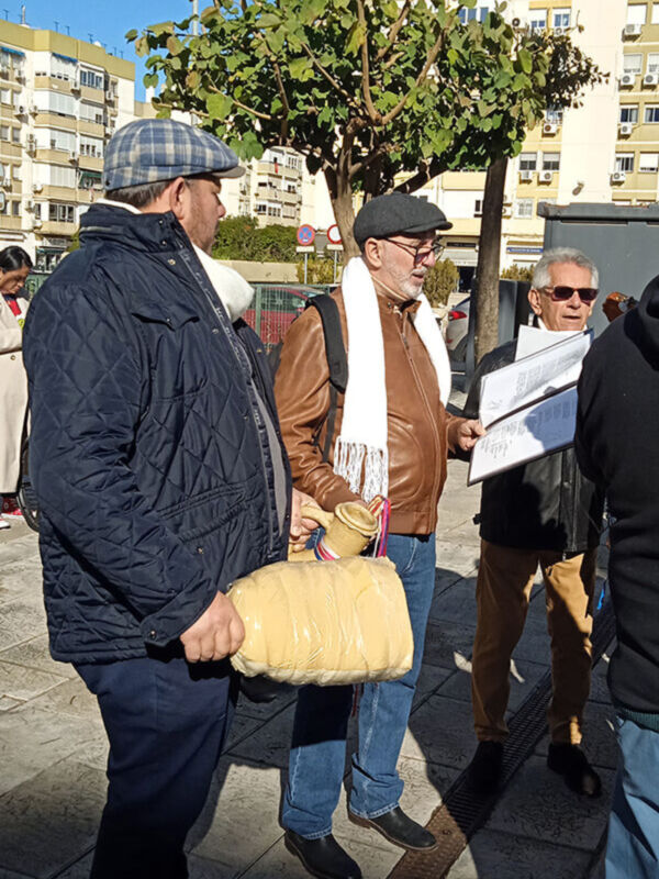 Una calle con gente caminando, algunos con gorras y chaquetas. Un hombre tiene una botella amarilla y un papel en la mano. Fondo de edificios, árboles y cielo azul claro.