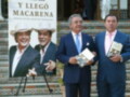 A tres hombres en trajes, uno de ellos sostiene un libro. El fondo es una pared con azulejos y una ventana con persianas. La imagen parece ser de un evento o presentación relacionada con la cultura o el arte, posiblemente en una ubicación tradicional.