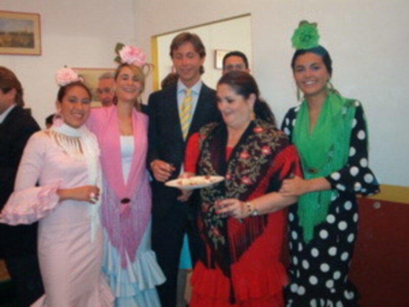 Una escena festiva donde una mujer en traje de flamenca ofrece un plato a una persona en traje formal, mientras otros individuos observan la escena.
