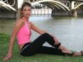 Una mujer con una camiseta rosa y pantalones negros se sienta en la hierba cerca de un puente sobre el río. La imagen muestra una escena relajada y relajante en un entorno urbano con agua y estructuras arquitectónicas.
