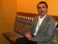 Un hombre con barba y bigote, vistiendo una chaqueta gris claro y camisa blanca, sentado en un banco con patrones de madera. La pared al fondo es amarilla y el banco tiene un tapizado con motivos de árboles.