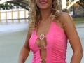 A una mujer con cabello largo y rizado, vestida con un top rosa brillante que tiene detalles metálicos. Está posando frente a un puente y un río, con una expresión sonriente.