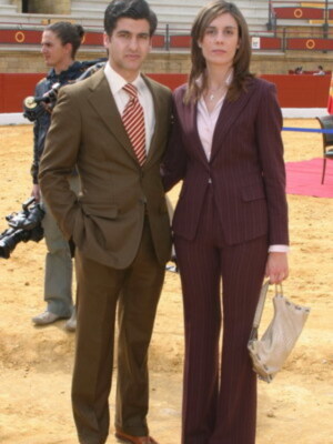 Dos personas posan en un estadio, uno con traje marrón y corbata roja, la otra con traje de chaqueta y pantalones a rayas. La imagen muestra una escena formal en un lugar público con gradas y un área de juego al fondo.