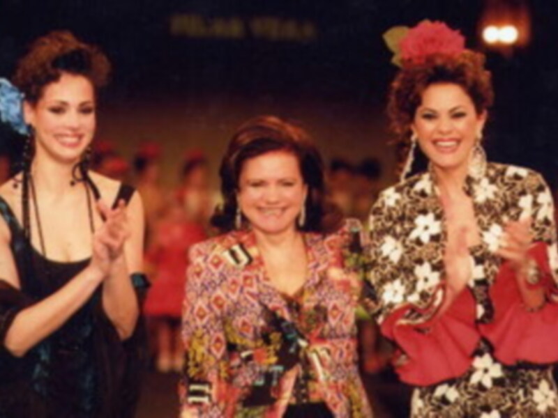 Tres mujeres en trajes tradicionales mexicanos, posando y aplaudiendo en un escenario nocturno.