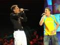 Dos cantantes en escenario, uno con camisa negra y pantalones blancos, el otro con camiseta amarilla y jeans.