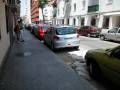 Un niño y un hombre caminan por una calle adoquinada, rodeados de coches estacionados frente a edificios de varios pisos. La escena es típica de un día laborable en una ciudad con edificios modernos y tradicionales.