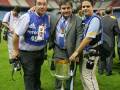 Tres hombres posan con un trofeo en un estadio iluminado, uno de ellos sostiene una cámara.