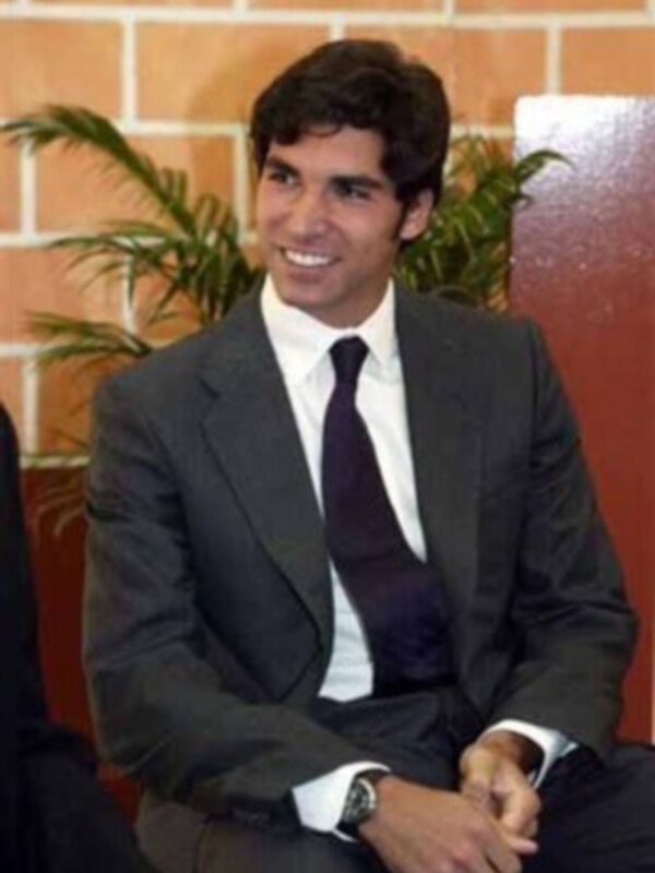 Persona en traje y corbata sonriendo, sentado en un entorno con pared de ladrillo y planta de fondo.