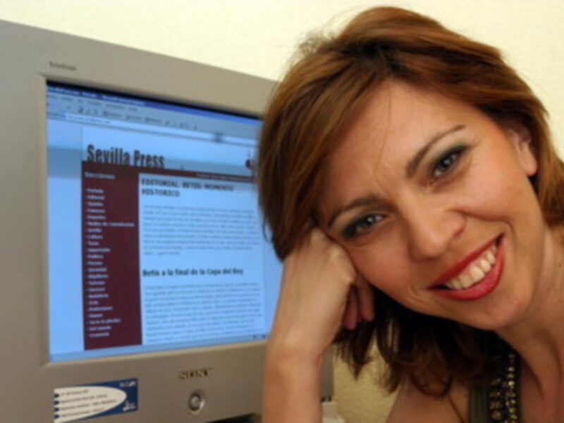 Una mujer sonríe frente a un monitor de computadora que muestra el sitio web de 'Sevilla Press'.