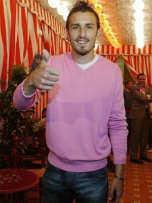 Un hombre en una camiseta rosa y pantalones vaqueros da un gesto de aprobación con su dedo en el aire. La imagen está ambientada en un ambiente festivo, con una decoración de cortinas rojas y blancas al fondo.