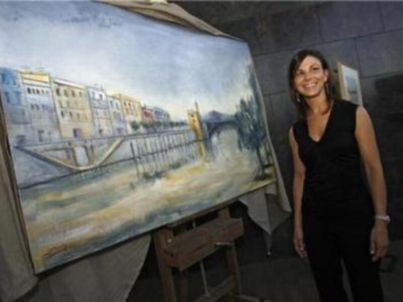 Artista posa junto a su obra acuarelista de un paisaje urbano con un puente y edificios históricos.