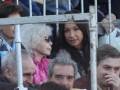 Una mujer con cabello blanco y gafas, sentada entre otros espectadores en un estadio.