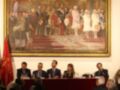 Un retrato histórico enmarcado de una reunión formal, con varias personas sentadas al frente y otros observando desde atrás. La escena parece ser de una reunión oficial o ceremonia importante, con vestimentas formales y un ambiente formal.