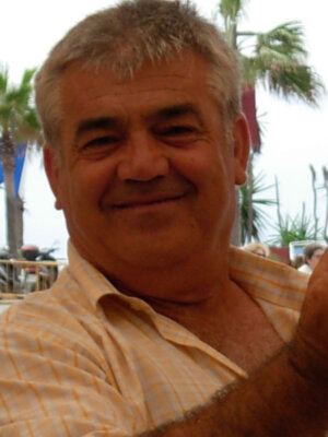 Un hombre con cabello gris y barba, sonriendo mientras toma una selfie en un ambiente al aire libre con palmeras y cielo despejado.