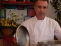 Un chef en uniforme blanco sostiene una sartén metálica. Detrás de él, se puede ver un escritorio con una pintura que dice "La Cocina de García". Hay flores y un recipiente en el escritorio. A la derecha, hay una planta con hojas verdes.