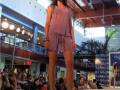 Una modelo camina por la pasarela en un evento de moda, vestida con un vestido rosa y sandalias. La audiencia observa desde los asientos alrededor de la pasarela, iluminada por luces y decoración.