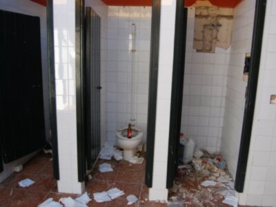 Bañera desgastada con baldosas rotas, piso rojo y puertas abiertas.