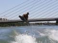 Un hombre realiza un salto de wakeboard en el agua, con una vista de la ciudad y un puente en el fondo.