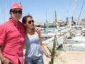 Dos personas posan en un muelle con barcos y banderas al fondo. La persona de la izquierda lleva una camisa roja, gafas y sombrero. La persona de la derecha usa un top azul claro y gafas. Ambos sonríen para la foto.