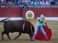 Un torero con un chaleco colorido y una capa roja se enfrenta a un toro negro en el centro de la plaza de toros. El público, vestido con camisetas rojas y azules, observa la escena desde la grada.