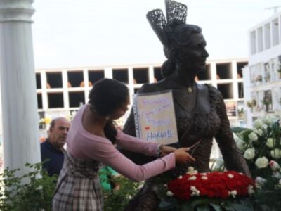 Una mujer coloca flores en una estatua de un hombre vestido con un sombrero. El fondo muestra un edificio con criptas y flores en las ventanas.