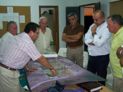 Un grupo de hombres discuten sobre un mapa colocado en una mesa cubierta con un mantel. La sala tiene un pizarrón de pared y una puerta abierta que muestra una vista exterior.