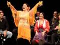 Una mujer en traje de flamenco con un vestido amarillo y flecos, levanta las manos en un gesto de danza. Detrás de ella, una mujer con vestido rojo y falda azul se sienta, mientras un músico toca el saxofón. En la orilla izquierda, dos hombres observan la actuación.