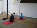 Tres personas sentadas en esterillas rojas en un espacio al aire libre, posiblemente practicando yoga.