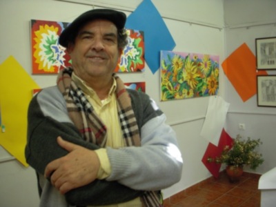 Un hombre sonriente con una bufanda y una camisa de manga larga posa frente a varias pinturas colgadas en la pared. La imagen muestra un ambiente artístico con diversos cuadros de flores y formas geométricas.