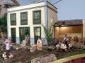 Un pequeño pueblo en miniatura con una casa blanca y verde, un jardín con figuras de personas sentadas y un niño en la entrada.