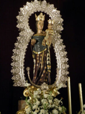 Estatua de una Virgen con un niño en sus brazos, rodeada por flores y adornos dorados.