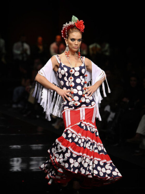 Modela desfilando en un evento de moda con un vestido flamenco tradicional de colores rojo, blanco y negro, adornado con detalles de flecos y flores en la cabeza.